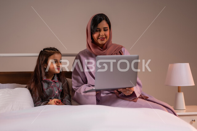 متابعة الدروس أونلاين، تحمل المسؤولية والعناية بالابناء، مفهوم التعلم عن بعد باستخدام الأجهزة التقنية الحديثة والتكنولوجيا، الأسرة السعودية ودورها في تربية إنشاء اجيال المستقبل، بورتريه لامرأة عربية خليجية سعودية محجبة تقوم بمساعدة ابنتها في دروسها، خلفية ملونة