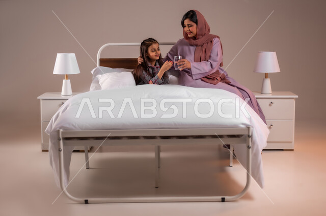 الاهتمام بصحة الابناء، المداومة على شرب المياه، المحافظة على سلامة الجهاز المناعي للجسم، الشعور بالعطش والجفاف، بورتريه لامرأة عربية خليجية سعودية تجلس على السرير تقوم بتقديم كوب ماء زجاجي لابنتها، خلفية ملونة