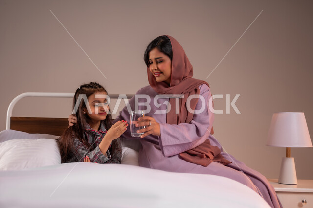 المداومة على شرب المياه، المحافظة على سلامة الجهاز المناعي للجسم، الشعور بالعطش والجفاف، بورتريه لامرأة عربية خليجية سعودية تجلس على السرير تقوم بتقديم كوب ماء زجاجي لابنتها، الاهتمام بصحة الابناء، خلفية ملونة