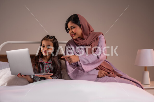 متابعة الدروس أونلاين، تحمل المسؤولية والعناية بالابناء، مفهوم التعلم عن بعد باستخدام الأجهزة التقنية الحديثة والتكنولوجيا، الأسرة السعودية ودورها في تربية إنشاء اجيال المستقبل، بورتريه لامرأة عربية خليجية سعودية محجبة تقوم بمساعدة ابنتها في دروسها، خلفية ملونة