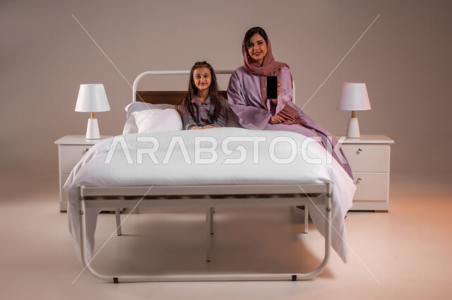 Looking at camera with happy gestures, showing blank black screen, development of technology and modern technical devices, portrait of smiling Saudi Arabian Gulf mother holding mobile phone in hand, little girl sitting on comfortable bed, concept of love and family bonding, colorful background