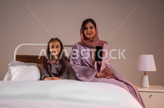 Blank black screen, looking at camera with happy gestures, development of technology and modern technical devices, portrait of smiling Saudi Arabian Gulf mother holding mobile phone in hand, little girl sitting on comfortable bed, concept of love and family bonding, colorful background