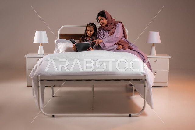 قضاء وقت ممتع مع الابنه، مفهوم التربيه القائمه على الحب والحنان، المشاركه بقراءه القصص المفيده مع الأبناء قبل النوم، بورتريه لامرأة عربية خليجية سعودية تقرأ الروايات المسليه لابنتها، خلفية ملونة