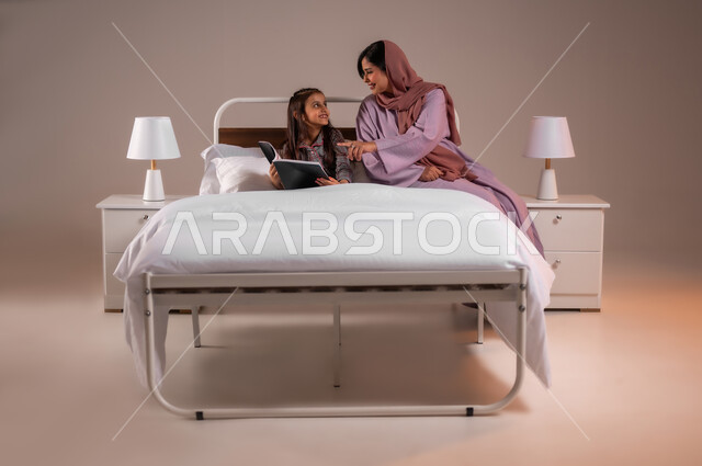 مفهوم التربيه القائمه على الحب والحنان، المشاركه بقراءه القصص المفيده مع الأبناء قبل النوم، بورتريه لامرأة عربية خليجية سعودية تقرأ الروايات المسليه لابنتها، قضاء وقت ممتع مع الأبنه قبل النوم، خلفية ملونة