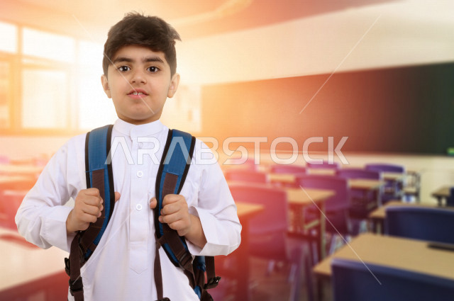 طالب مدرسي عربي خليجي سعودي يقف في الصف المدرسي  ، بلباس سعودي خليجي تقليدي ، يرتدي حقيبة ظهر مدرسية ، يستعد للفصل الدراسي الجديد ، فصل دراسي تعليمي ، الاستعداد للدروس والواجبات ، الاستعداد للمدارس ، مع خلفية فصل دراسي