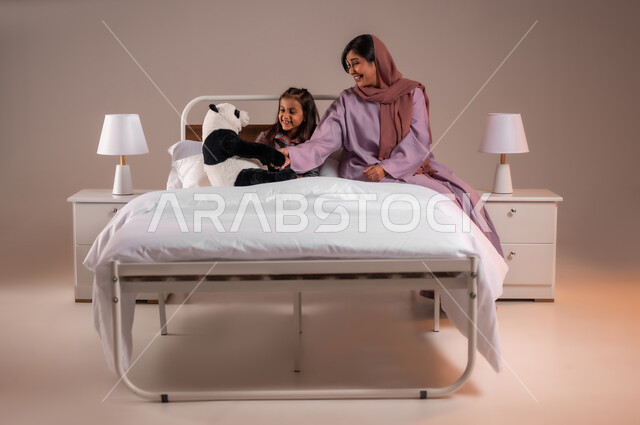 ممارسة الأنشطة الترفيهية المسلية، تحمل المسؤولية والعناية بالابناء، تقوية رابط الأمومة بين الأم وطفلتها، قضاء أوقات ممتعة مع الاولاد، بورتريه لامرأة عربية خليجية سعودية تقوم باللعب بدمية الدب مع ابنتها، خلفية ملونة