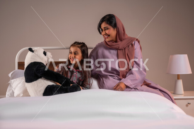 تحمل المسؤولية والعناية بالابناء، تقوية رابط الأمومة بين الأم وطفلتها، قضاء أوقات ممتعة مع الاولاد، ممارسة الأنشطة الترفيهية المسلية، بورتريه لامرأة عربية خليجية سعودية تقوم باللعب بدمية الدب مع ابنتها، خلفية ملونة