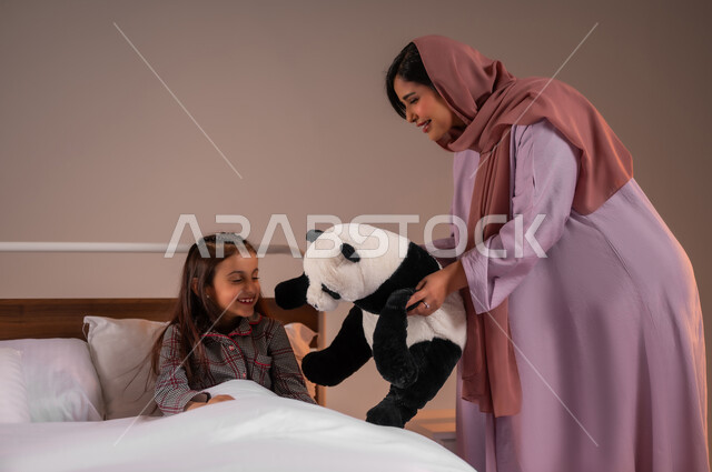 تقوية رابط الأمومة بين الأم وطفلتها، تحمل المسؤولية والعناية بالابناء، قضاء أوقات ممتعة مع الاولاد، ممارسة الأنشطة الترفيهية المسلية، بورتريه لامرأة عربية خليجية سعودية تقوم باللعب بدمية الدب مع ابنتها، خلفية ملونة