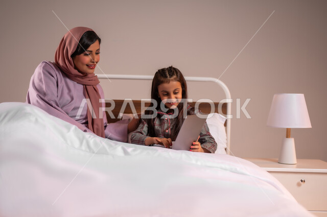 الأسرة السعودية ودورها في تربية إنشاء اجيال المستقبل، مفهوم التعلم عن بعد باستخدام الأجهزة التقنية الحديثة والتكنولوجيا، تحمل المسؤولية والعناية بالابناء، متابعة الدروس أونلاين، بورتريه لامرأة عربية خليجية سعودية محجبة تقوم بمساعدة ابنتها في دروسها، خلفية ملونة