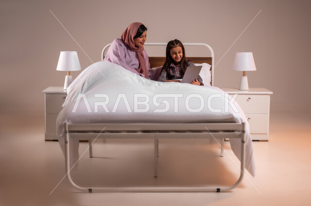 مفهوم التعلم عن بعد باستخدام الأجهزة التقنية الحديثة والتكنولوجيا، تحمل المسؤولية والعناية بالابناء، متابعة الدروس أونلاين، الأسرة السعودية ودورها في تربية إنشاء اجيال المستقبل، بورتريه لامرأة عربية خليجية سعودية محجبة تقوم بمساعدة ابنتها في دروسها، خلفية ملونة