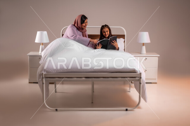 مفهوم التربيه القائمه على الحب والحنان، المشاركه بقراءه القصص المفيده مع الأبناء قبل النوم، بورتريه لامرأة عربية خليجية سعودية تقرأ الروايات المسليه لابنتها، قضاء وقت ممتع مع الأبنه قبل النوم، خلفية ملونة