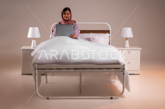Concept of advanced modern technology and techniques, accomplishing work tasks remotely, portrait of a smiling Saudi Arabian Gulf woman sitting in a comfortable bed using a laptop, making video calls over the internet, colorful background