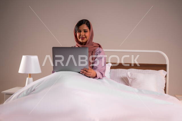 Completing work tasks remotely, concept of advanced modern technology and techniques, portrait of a smiling Saudi Arabian Gulf woman sitting in a comfortable bed using a laptop, making video calls over the internet, colorful background