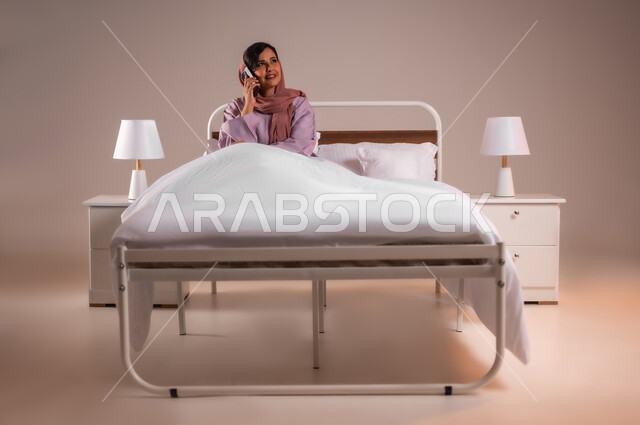 التواصل مع الأهل والأصدقاء باستخدام الاجهزة التقنية الحديثة، محادثات وحوارات ودية، ايماءات السعادة و السرور، بورتريه لامراة عربية خليجية سعودية محجبة مبتسمة تجلس على سرير مريح تحمل بيدها الهاتف المحمول تقوم باجراء مكالمة هاتفية، خلفية ملونة