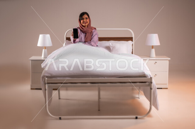 Showing a blank black screen, looking at the camera with happy gestures, development of technology and modern technical devices, portrait of a smiling Saudi Arabian Gulf woman wearing an abaya sitting on a comfortable bed holding a mobile phone in her hand, colorful background