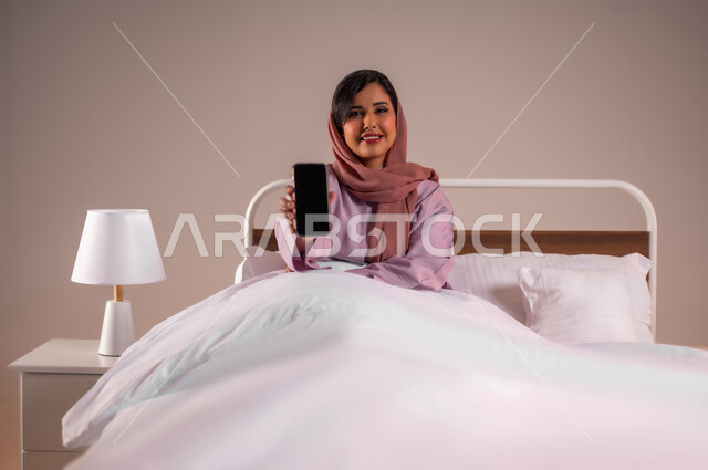 Development of technology and modern technical devices, displaying a blank black screen, looking at the camera with gestures of happiness, portrait of a smiling Saudi Arabian Gulf woman wearing an abaya sitting on a comfortable bed holding a mobile phone in her hand, colorful background