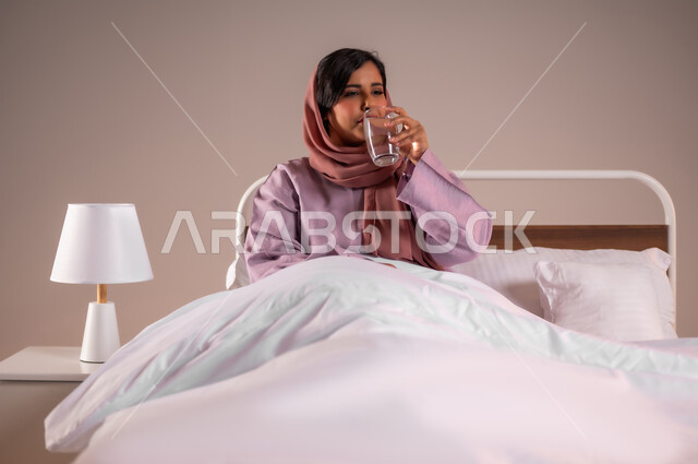 المحافظة على سلامة الجهاز المناعي للجسم، المداومة على شرب المياه، الشعور بالعطش والجفاف، بورتريه لامرأة عربية خليجية سعودية محجبة تجلس على سرير مريح تحمل بيدها كوب زجاجي تقوم بشرب الماء، خلفية ملونة