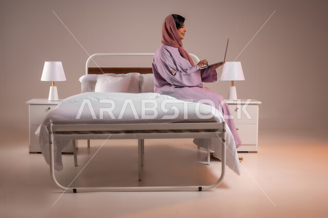 Concept of advanced modern technology and techniques, accomplishing work tasks remotely, portrait of a smiling Saudi Arabian Gulf woman sitting in a comfortable bed using a laptop, making video calls over the internet, colorful background