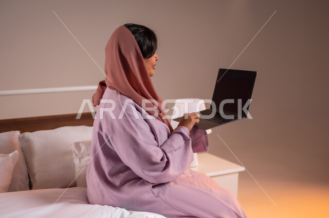 Completing work tasks remotely, concept of advanced modern technology and techniques, portrait of a smiling Saudi Arabian Gulf woman sitting in a comfortable bed using a laptop, making video calls over the internet, colorful background