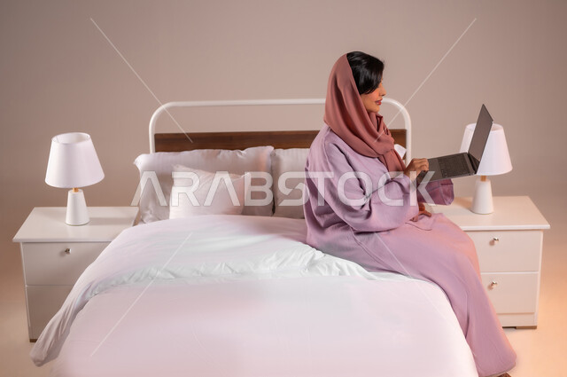 إنجاز مهام العمل عن بعد، مفهوم التقنيات والتكنولوجيا الحديثة المتطورة، بورتريه لامرأة عربية خليجية سعودية مبتسمة تجلس في سرير مريح تقوم باستخدام جهاز الحاسوب المحمول، إجراء مكالمات فيديو عبر الانترنت، خلفية ملونة