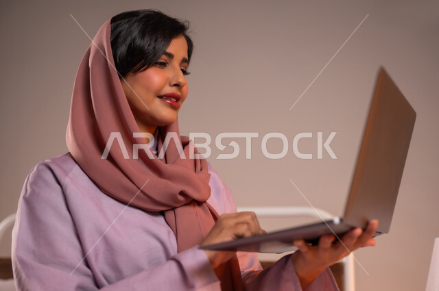 إجراء مكالمات فيديو عبر الانترنت، إنجاز مهام العمل عن بعد، مفهوم التقنيات والتكنولوجيا الحديثة المتطورة، بورتريه مقرب لامرأة عربية خليجية سعودية مبتسمة تجلس في سرير مريح تقوم باستخدام جهاز الحاسوب المحمول، خلفية ملونة