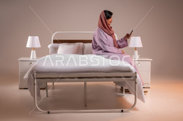 استخدام الاجهزة التقنية الحديثة والمتطورة، الدردشه مع الأهل والأصدقاء، تصفح تطبيقات وبرامج التواصل الاجتماعي، إيماءات السرور والسعادة، بورتريه لسيدة عربية خليجية سعودية محجبة مبتسمة تجلس في سرير مريح تمسك بيدها الهاتف المحمول، خلفية ملونة