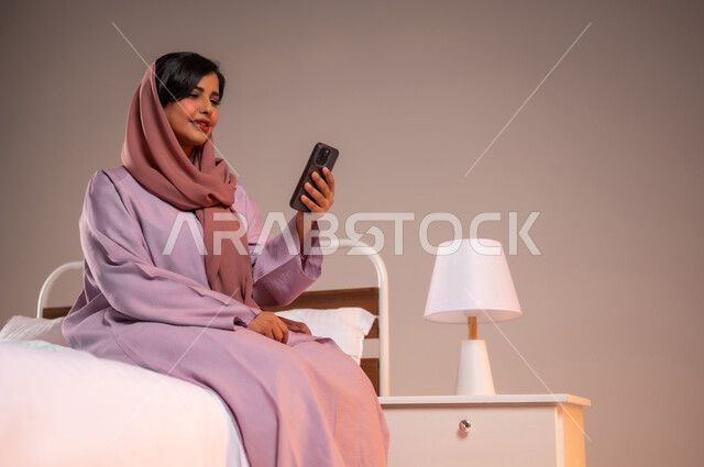 الدردشه مع الأهل والأصدقاء، تصفح تطبيقات وبرامج التواصل الاجتماعي، إيماءات السرور والسعادة، بورتريه لسيدة عربية خليجية سعودية محجبة مبتسمة تجلس في سرير مريح تمسك بيدها الهاتف المحمول، استخدام الاجهزة التقنية الحديثة والمتطورة، خلفية ملونة