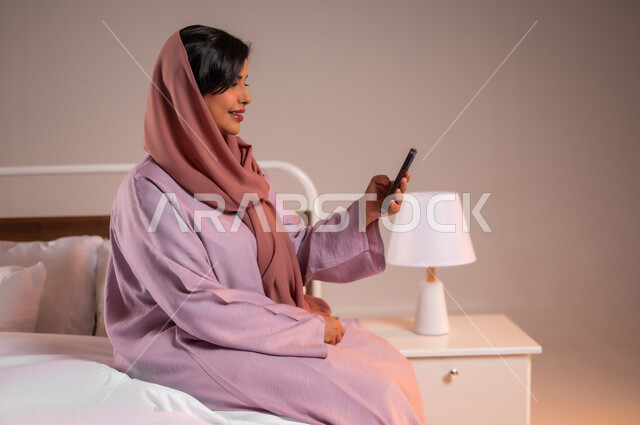 Browsing social media applications and programs, gestures of pleasure and happiness, chatting with family and friends, portrait of a smiling veiled Saudi Arabian Gulf woman sitting in a comfortable bed holding a mobile phone in her hand, using modern and advanced technical devices, colorful background