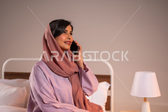 ايماءات السعادة و السرور، محادثات وحوارات ودية، التواصل مع الأهل والأصدقاء باستخدام الاجهزة التقنية الحديثة، بورتريه مقرب لامراة عربية خليجية سعودية محجبة مبتسمة تجلس على سرير مريح تحمل بيدها الهاتف المحمول تقوم باجراء مكالمة هاتفية، خلفية ملونة