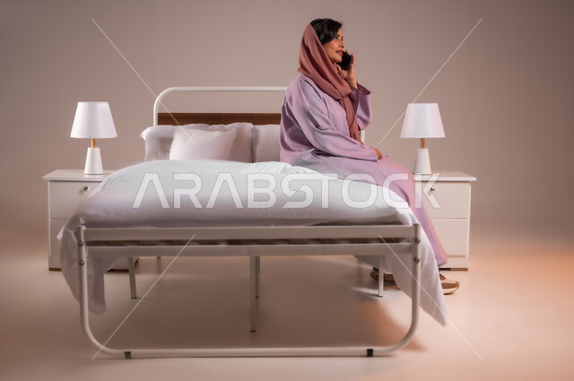 محادثات وحوارات ودية، التواصل مع الأهل والأصدقاء باستخدام الاجهزة التقنية الحديثة، ايماءات السعادة و السرور، بورتريه لامراة عربية خليجية سعودية محجبة مبتسمة تجلس على سرير مريح تحمل بيدها الهاتف المحمول تقوم باجراء مكالمة هاتفية، خلفية ملونة