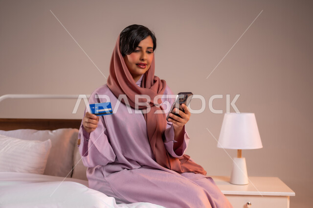 التكنولوجيا الحديثة والتقنيات المتطورة، استخدام البطاقة البنكية الالكترونية، إمرأة عربية سعودية خليجية محجبة مبتسمة تجلس على سرير مريح تقوم بالتسوق باستعمال الهاتف المحمول، إيماءات السعادة والسرور، مفهوم الدفع عن بعد عبر تطبيقات الجوال، خلفية ملونة