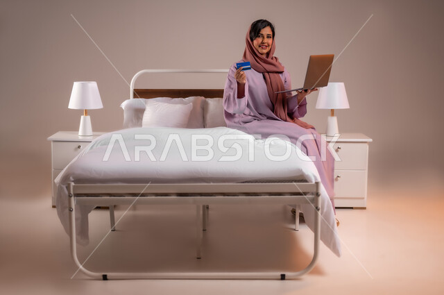 The concept of remote payment via laptop applications, using an electronic bank card, a portrait of a smiling veiled Saudi Arabian Gulf woman sitting on a bed shopping via a laptop, using modern technical devices, a colorful background