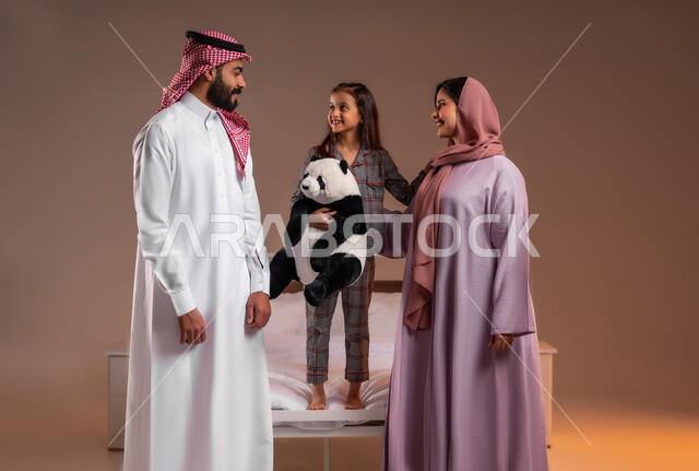 The concept of family love and tenderness, getting ready for sleep, a portrait of a Saudi Arabian Gulf girl wearing pajamas, holding a bear-shaped toy in her hand, standing on the bed next to her parents, spending fun family times, colorful background
