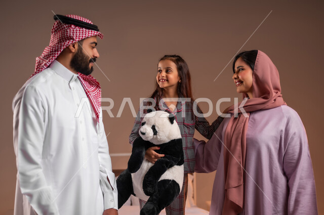 الاستعداد والتجهز للنوم، بورتريه لفتاة عربية خليجية سعودية ترتدي البيجامة تحمل بيدها لعبة على شكل دب تقف على السرير بجانب والديها، مفهوم الحب والحنان الاسري، قضاء أوقات عائلية ممتعة، خلفية ملونة