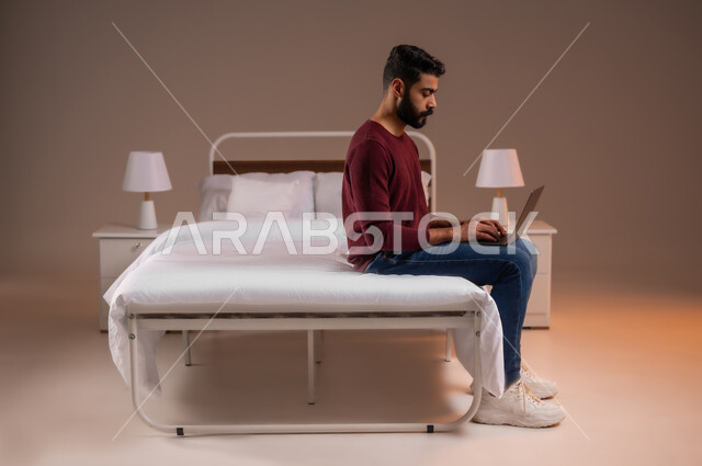 إستخدام الحاسوب المحمول لإنجاز المهام، عقد إجتماع عمل عبر الإنترنت، تصفح برامج التواصل الاجتماعي, بورتريه لرجل عربي خليجي سعودي يرتدي الزي الكاجوال يحمل جهاز اللابتوب يجلس على سريره في غرفة النوم، تطور التكنولوجيا والأجهزة التقنية، صورة لكامل الجسم، خلفية ملونة
