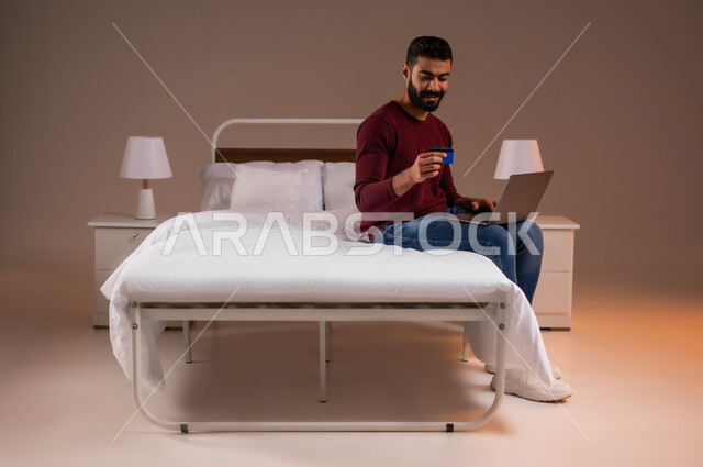 مفهوم الدفع والتسديد عن بعد عبر تطبيقات اللابتوب، استخدام البطاقة البنكية الالكترونية، تطور التكنولوجيا والأجهزة التقنية، بورتريه لرجل عربي سعودي خليجي يرتدي الزي الكاجوال يجلس على سرير مريح يعمل على جهاز الحاسوب المحمول، صورة لكامل الجسم، خلفية ملونة