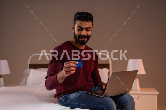 استخدام البطاقة البنكية الالكترونية، مفهوم الدفع والتسديد عن بعد عبر تطبيقات اللابتوب، تطور التكنولوجيا والأجهزة التقنية، بورتريه لرجل عربي سعودي خليجي يرتدي الزي الكاجوال يجلس على سرير مريح يعمل على جهاز الحاسوب المحمول، خلفية ملونة
