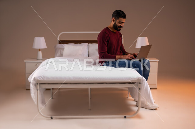 إستخدام الحاسوب المحمول لإنجاز المهام، عقد إجتماع عمل عبر الإنترنت، تصفح برامج التواصل الاجتماعي, بورتريه لرجل عربي خليجي سعودي يرتدي الزي الكاجوال يحمل جهاز اللابتوب يجلس على سريره في غرفة النوم، تطور التكنولوجيا والأجهزة التقنية، صورة لكامل الجسم، خلفية ملونة