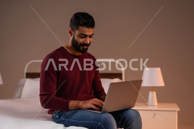 Holding a business meeting online, browsing social media, using laptop to complete tasks, portrait of a Saudi Arabian Gulf man in casual wear holding a laptop sitting on his bed in bedroom, development of technology and technical devices, colorful background
