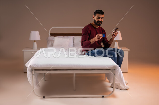 استخدام البطاقة البنكية الالكترونية، التسوق الالكتروني عبر الانترنت، مفهوم الدفع والتسديد عن بعد عبر تطبيقات الجوال، بورتريه لرجل عربي سعودي خليجي يرتدي الزي الكاجوال يجلس على سرير مريح يحمل بيده الهاتف المحمول، صورة لكامل الجسم، خلفية ملونة