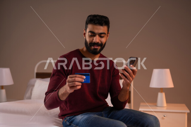 التسوق الالكتروني عبر الانترنت، مفهوم الدفع والتسديد عن بعد عبر تطبيقات الجوال، استخدام البطاقة البنكية الالكترونية، بورتريه لرجل عربي سعودي خليجي يرتدي الزي الكاجوال يجلس على سرير مريح يحمل بيده الهاتف المحمول، خلفية ملونة