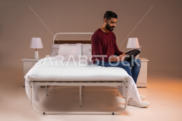 هواية المطالعة والاستكشاف، قضاء وقت الفراغ بالتعلم والثقافة، مفهوم التثقيف والبحث العلمي، بورتريه لشاب عربي خليجي سعودي مبتسم يرتدي الزي الكاجوال يجلس على سرير مريح يقوم قراءة كتاب, خلفية ملونة