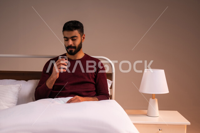 المداومة على شرب المياه، المحافظة على سلامة الجهاز المناعي للجسم، الشعور بالعطش والجفاف، بورتريه لرجل عربي خليجي سعودي يرتدي الزي الكاجوال يستلقي على سرير مريح يحمل بيده كوب زجاجي يقوم بشرب الماء، خلفية ملونة