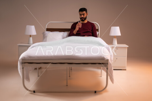 المحافظة على سلامة الجهاز المناعي للجسم، المداومة على شرب المياه، الشعور بالعطش والجفاف، بورتريه لرجل عربي خليجي سعودي يرتدي الزي الكاجوال يستلقي على سرير مريح يحمل بيده كوب زجاجي يقوم بشرب الماء، خلفية ملونة