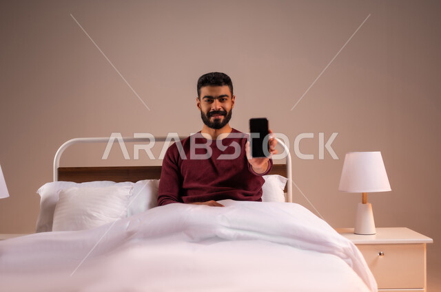 النظر الى الكاميرا بايماءات السعادة، عرض شاشة سوداء فارغة، تطور التكنولوجيا والأجهزة التقنية الحديثة، بورتريه لرجل عربي سعودي خليجي مبتسم يرتدي الزي الكاجوال يستلقي على سرير مريح يحمل بيده الهاتف المحمول، خلفية ملونة