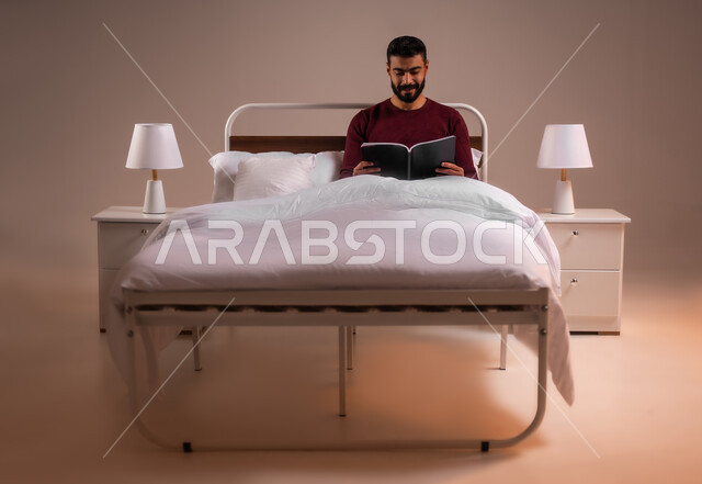 قضاء وقت الفراغ بالتعلم والثقافة، هواية المطالعة والاستكشاف، مفهوم التثقيف والبحث العلمي، بورتريه لشاب عربي خليجي سعودي مبتسم يرتدي الزي الكاجوال مستلقي على سرير مريح يقوم قراءة كتاب, خلفية ملونة