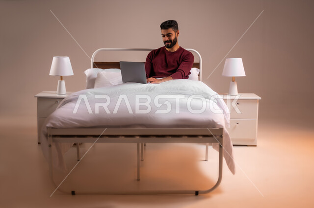 عقد إجتماع عمل عبر الإنترنت، تصفح برامج التواصل الاجتماعي, إستخدام الحاسوب المحمول لإنجاز المهام، بورتريه لرجل عربي خليجي سعودي يرتدي الزي الكاجوال يحمل جهاز اللابتوب يستلقي على سريره في غرفة النوم، تطور التكنولوجيا والأجهزة التقنية، خلفية ملونة