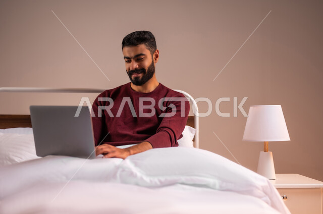 عقد إجتماع عمل عبر الإنترنت، تصفح برامج التواصل الاجتماعي, إستخدام الحاسوب المحمول لإنجاز المهام، بورتريه لرجل عربي خليجي سعودي يرتدي الزي الكاجوال يحمل جهاز اللابتوب يستلقي على سريره في غرفة النوم، تطور التكنولوجيا والأجهزة التقنية، خلفية ملونة