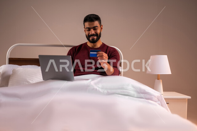 استخدام البطاقة البنكية الالكترونية، تطور التكنولوجيا والأجهزة التقنية، مفهوم الدفع والتسديد عن بعد عبر تطبيقات اللابتوب، بورتريه لرجل عربي سعودي خليجي يرتدي الزي الكاجوال مستلقي على سرير مريح يعمل على جهاز الحاسوب المحمول، خلفية ملونة
