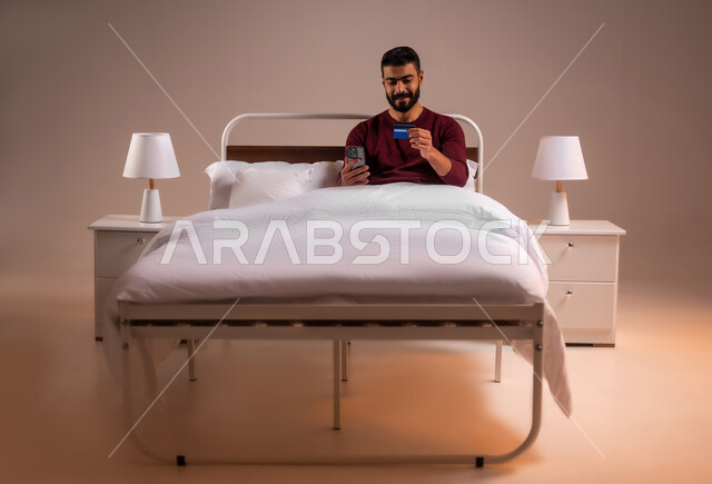 التسوق الالكتروني عبر الانترنت، مفهوم الدفع والتسديد عن بعد عبر تطبيقات الجوال، استخدام البطاقة البنكية الالكترونية، بورتريه لرجل عربي سعودي خليجي يرتدي ازي الكاجوال مستلقي على سرير مريح يحمل الهاتف المحمول، خلفية ملونة